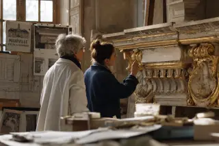 Two conservators of different generations examine details on an ornate cornice in a softly lit, authentic restoration workshop, surrounded by old plans and reference photos, in an atmosphere of quiet concentration.