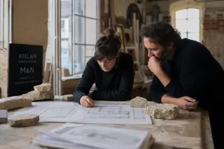Two architects in a softly lit Maxéville workshop examine stone samples and old plans on a wooden bench, with morning light highlighting their natural, focused expressions amid historic brick walls.