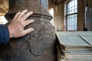 Hand touching engraved cast-iron column in old industrial building, morning light highlighting aged textures and archival blueprints nearby