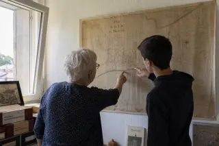 Elderly woman and teenage boy studying an old map together in a softly lit, authentic municipal archive room, conveying a quiet moment of shared curiosity about heritage.