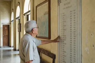Elderly man gently touching a marble plaque in a dim, historic municipal hallway with natural evening light and visible signs of age.