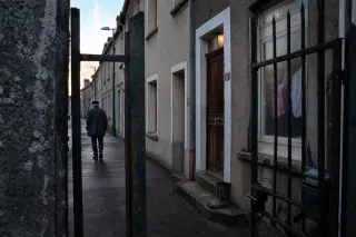 Perspective from behind a weathered gate, showing Maxéville workers’ houses at dawn with subtle signs of daily life and a solitary resident walking away in soft morning light.
