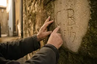 Hands of an older resident touch carved initials on a weathered stone wall in a narrow alley, soft afternoon light highlighting historical detail.