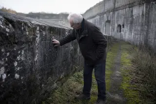 Elderly man examining lichen-covered engraving on the stone wall of Fort de Maxéville in soft morning light, surrounded by mossy path and historic architecture.
