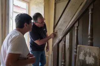 Two people closely examining handwritten notes on an old wooden staircase in a sunlit Maxéville house, authentic details and soft natural light emphasize the search for family history.