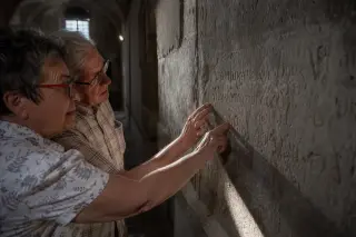 Two elderly locals examine ancient graffiti on a stone wall in a softly lit historic Maxéville corridor, highlighting tactile connection with heritage.